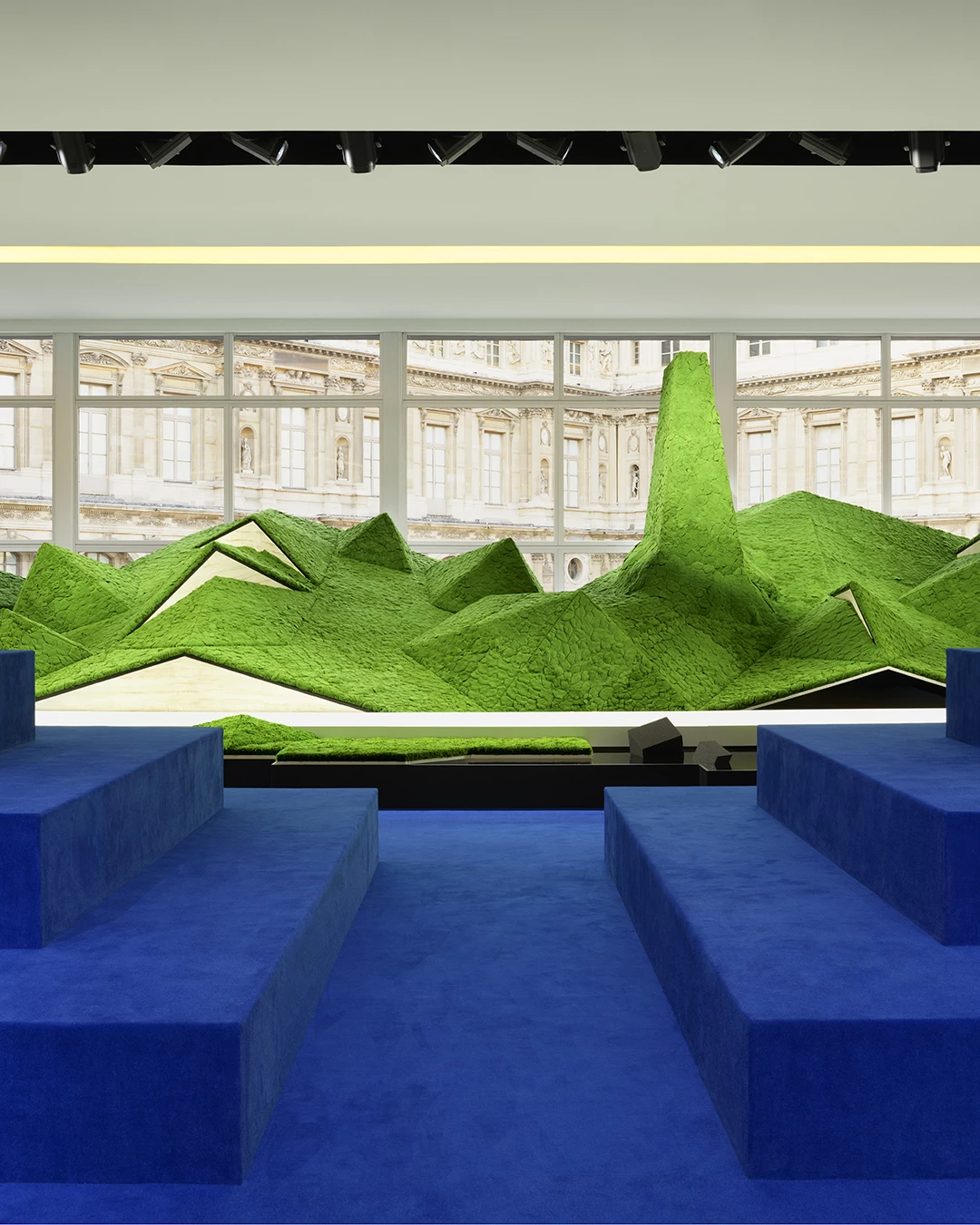 這一季 Louis Vuitton 依舊選在羅浮宮 Cour Carrée 方形庭院發表女裝系列，並找來《人生切割術》美術指導 Jeremy Hindle 操刀場景。走進秀場，你會彷彿走進虛實之間的空間，深淺不一的綠色地景、幾何起伏如山嵐的裝置、被置於舞台中央的賓客視角，打造的場景像是介於科幻地景，又好像是高山美景，而這其實是整場秀最重要的起點，品牌想敘述自然、旅行與未來科技如何重新交會，因此模特兒不是走在一條平坦秀道上，而像在穿越某種被重新構築的地貌，