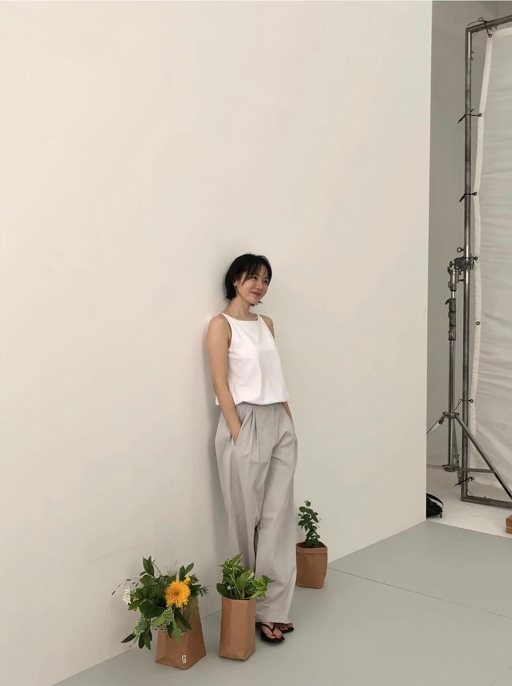 喜歡極簡俐落風,這套白背心配寬褲是通勤與日常的萬用公式。孫藝珍以一件白色船型領無袖上衣,搭配灰色打褶寬褲,微微將上衣紮進褲頭強調腰線,再以黑色細帶涼鞋收尾,整體乾淨中性又有鬆弛感。船型領修飾肩線、寬褲拉長比例,白配灰的低調配色更顯質感,是想穿得簡單卻不無聊的最佳範本。