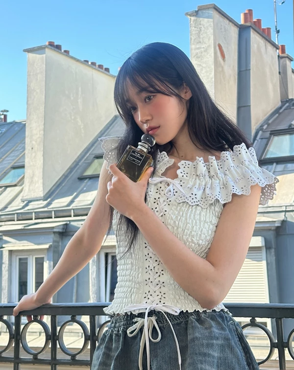 這張在歐洲陽台邊的側影，簡直是法式少女的完美化身！盧允瑞聰明地選擇了細緻的縐褶剪裁上衣，領口那一圈精緻的鏤空蕾絲荷葉邊，在陽光下泛著溫柔的光芒，不僅修飾了肩頸線條，更把少女的甜美與純淨拉滿，隨手拿著香水瓶就散發著迷人的鬆弛感。
