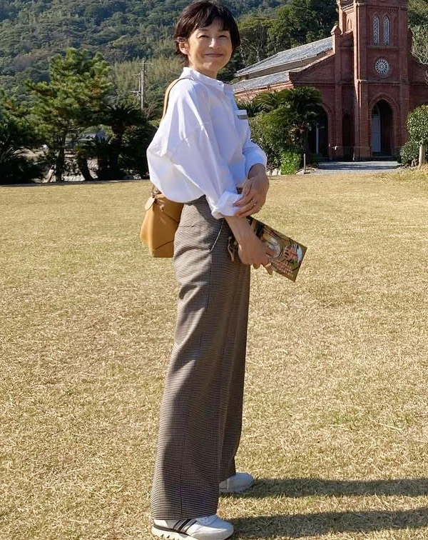 在藍天綠地襯托下,鈴木保奈美以寬鬆的廓形白襯衫,搭配格紋高腰寬褲與運動鞋,後背一個焦糖色皮革包,整個人顯得非常輕盈自在。一般人在穿基本款白襯衫時,千萬不要把釦子扣得死緊,或選太合身的版型。學她挑選落肩、帶有空氣感的寬鬆襯衫,將袖口隨意挽起,並把下擺紮進高腰褲中帶出好比例,配上一雙厚底白鞋,就能完美複製這份兼具舒適與文藝感的旅行逆齡感。