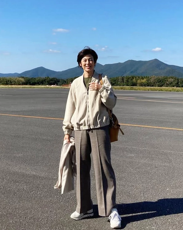 站在遼闊停機坪上的鈴木保奈美,換上了極具空氣感的米白色立領休閒夾克,下半身則混搭了一條垂墜感極佳的棕色格紋寬褲與乾淨白鞋。一般人穿運動風夾克時,很容易顯得太邋遢或像去運動,祕訣就在於「風格混搭」。學她用帶有正裝感的細格紋西褲來搭配輕便的廓形夾克,內搭則選擇低調的素色T,這種「半休閒、半正式」的組合,不僅穿起來舒適,更能展現出不經意、放鬆,且極度減齡的文藝旅人風範。