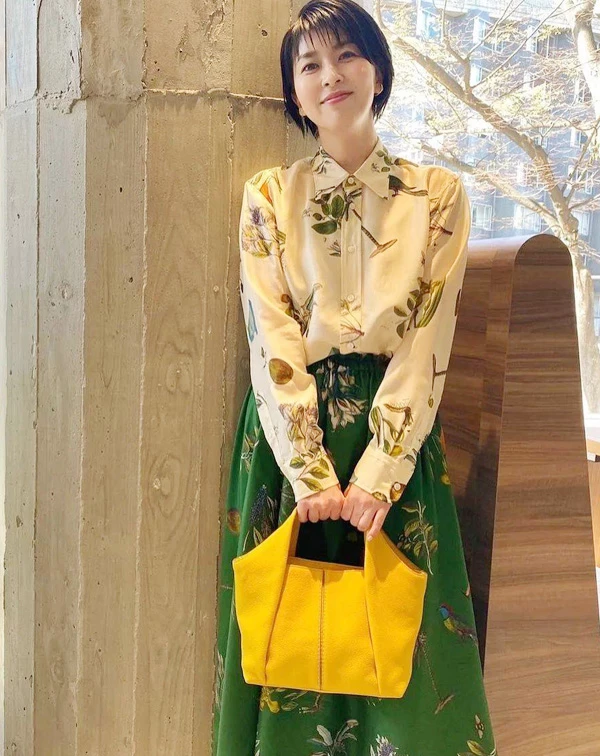 松隆子很適合這種帶點文藝感的穿搭。她用植物圖鑑印花襯衫搭配森林綠 A 字長裙,雖然上下身都有印花元素,但因為整體色調都落在綠色、米色和黃色這種偏自然的大地色系,所以看起來完全不會太花,反而有種很舒服的層次感。她把襯衫下擺紮進裙子裡,也讓整體比例變得更俐落、有精神。最亮眼的還是那款鮮黃Tod's色托特包,剛好替整體偏沉穩的配色多了一點活力,讓這套穿搭看起來既有氣質,又不會太成熟。