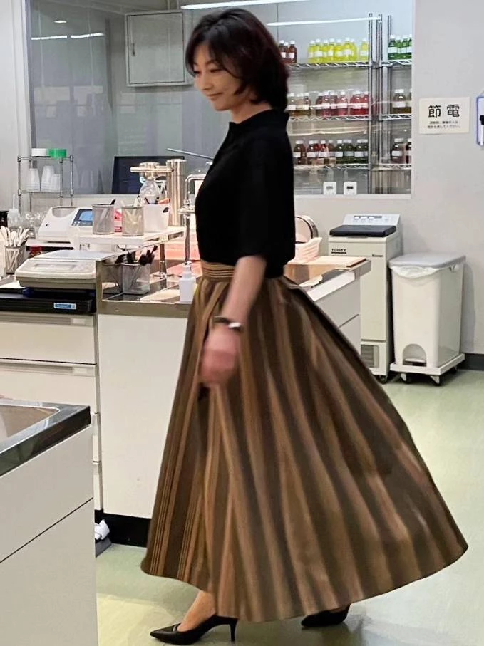 常盤貴子展示了最適合職場女性的俐落穿搭。她以一件設計簡約的黑色五分袖襯衫，搭配一條具有光澤感的棕色直條紋傘裙。將襯衫紮入裙中，利用裙子的垂墜感與寬鬆下襬，修飾下半身，整體比例極佳，展現出成熟女性從容不迫的自信美。