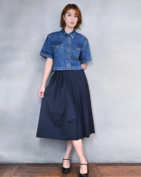 如何將「優雅傘裙」穿出率性又不失層次的高級質感？曾沛慈聰明地選用一件深藍色高腰百褶長裙，利用沉穩的色系與大面積的垂直褶皺，視覺上不僅顯瘦，更散發出一股知性的學院氣息。最巧妙的混搭技巧在於上半身，她沒有選擇傳統的襯衫，而是穿上一件短版短袖丹寧外套。外套的硬挺版型與金屬釦飾為溫柔的傘裙注入了「工裝感」的丹寧粗獷，展現出俐落與優雅共存的時尚態度。