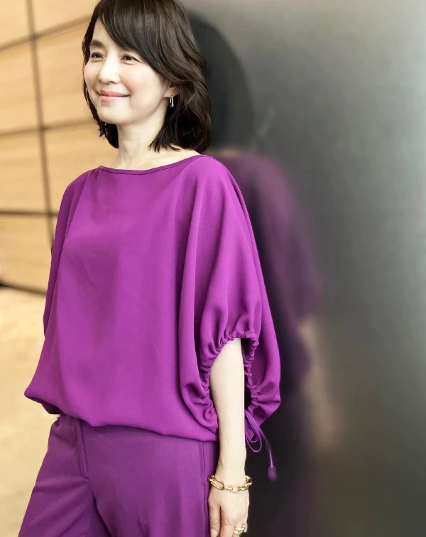 現年57歲的石田百合子，完美示範了如何透過服裝穿出年輕氣息。就像這套，她選擇了一身搶眼的亮紫色上衣與長褲。雖然顏色鮮豔大膽，但衣服本身簡約大方的剪裁與高質感的布料，讓整體造型顯得時髦而非過於前衛。上衣的蓬蓬袖與縮口袖口設計，則為熟女性魅力增添了一抹俏皮的年輕感。而這正是一個聰明的減齡穿搭技巧！在單品上運用一些帶有年輕感的元素，如：荷葉邊、縮口袖口等。