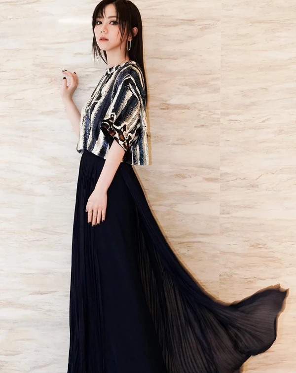 這套造型裡，鄧紫棋展現的是一種介於華麗與克制之間的清冷仙氣。她沒有選擇常見的飄逸洋裝輪廓，而是以短版圖騰上衣搭配落地黑色百褶長裙，透過鮮明對比，拉出俐落而充滿張力的比例線條。