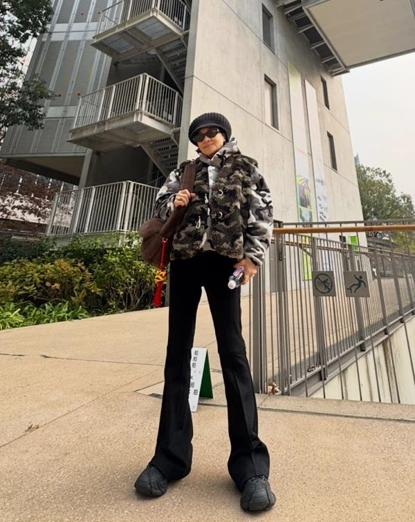 誰說70+不能挑戰層次感?她選用一件灰白迷彩連帽上衣,外搭一件具有厚實質感的深色刷毛背心,這種層疊穿法不僅保暖,更展現了街頭潮流中必備的份量感。下半身則配上極簡的黑色微喇叭長褲,完美拉長了腿部線條,讓整體比例看起來既高挑又輕盈。